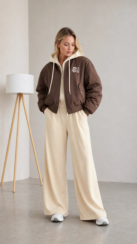 Front full view of woman wearing brown layered hooded bomber jacket with cream wide-leg pants