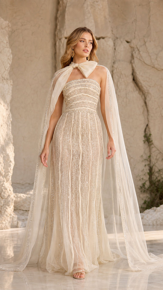 Front view of champagne strapless beaded maxi gown with flowing sheer cape and bow collar