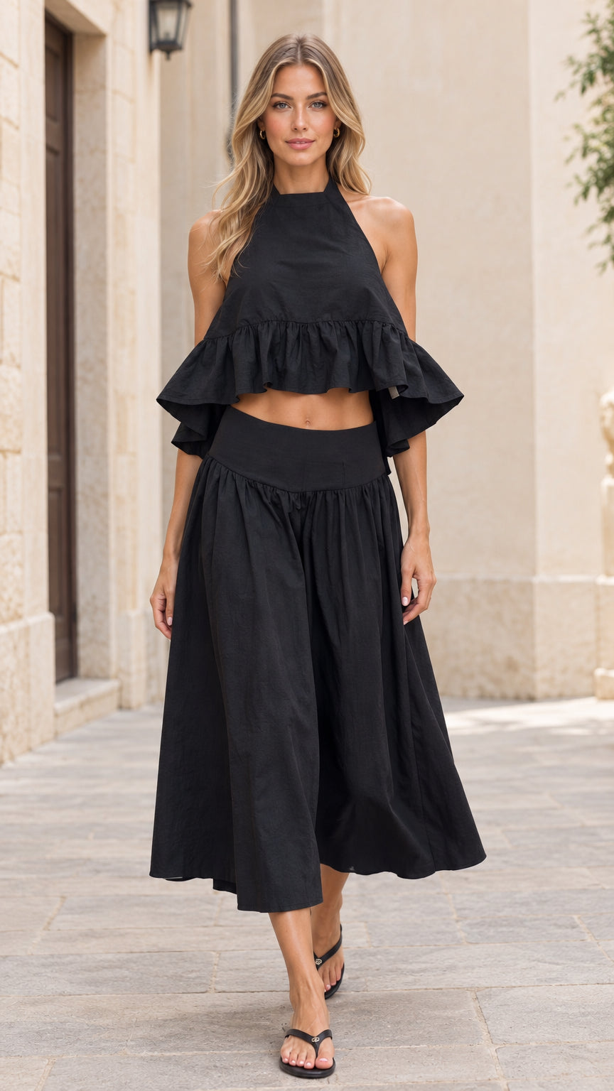 Front full-length view of model wearing black halter ruffle crop top and matching midi skirt set